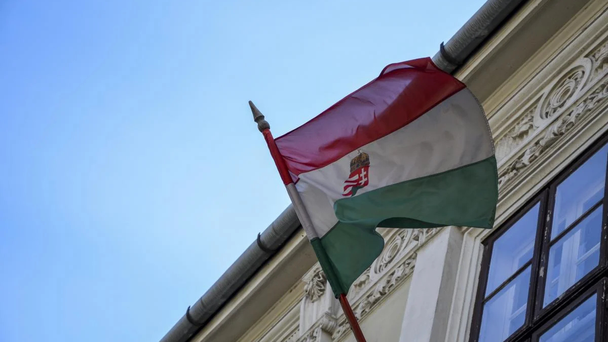 Hungarian flag waving on a flagpole beside an ornate building façade against a blue sky, showing red-white-green stripes and crest in the white stripe.