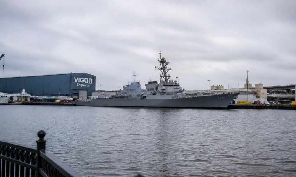 Gray naval destroyer moored at a pier with a blue VIGOR Shipyards building in the background.