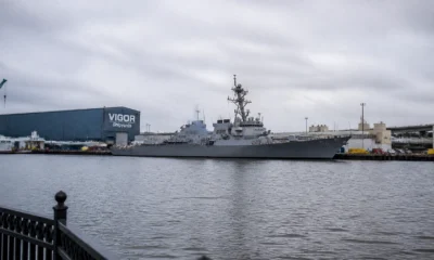 Gray naval destroyer moored at a pier with a blue VIGOR Shipyards building in the background.