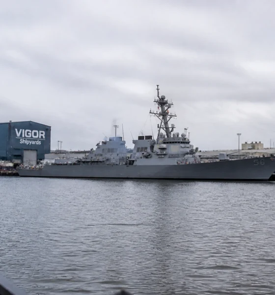 Gray naval destroyer moored at a pier with a blue VIGOR Shipyards building in the background.
