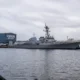 Gray naval destroyer moored at a pier with a blue VIGOR Shipyards building in the background.