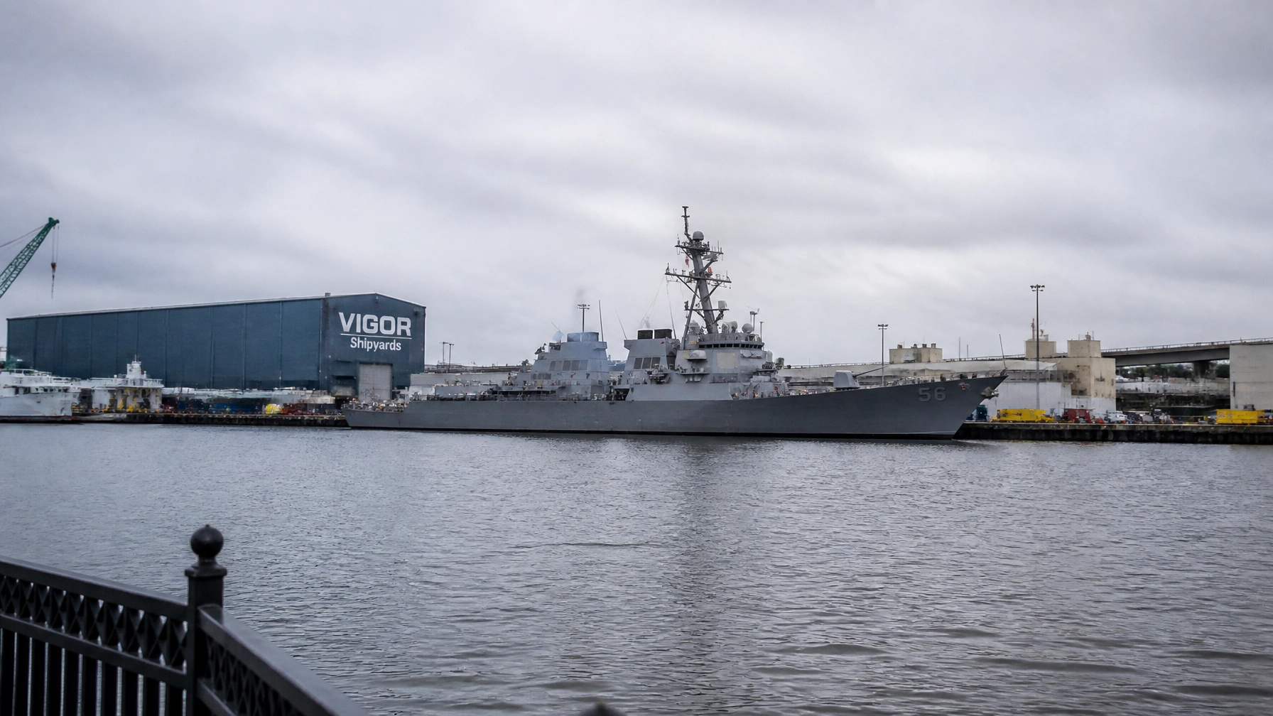 Gray naval destroyer moored at a pier with a blue VIGOR Shipyards building in the background.