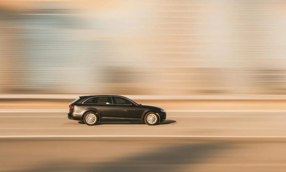 Dark station wagon speeding along a sunlit highway with motion blur.