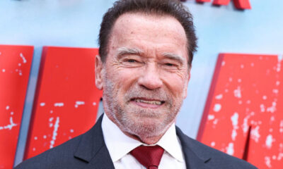 Smiling man in a dark suit and white shirt with a red tie, standing in front of a red backdrop.