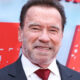 Smiling man in a dark suit and white shirt with a red tie, standing in front of a red backdrop.