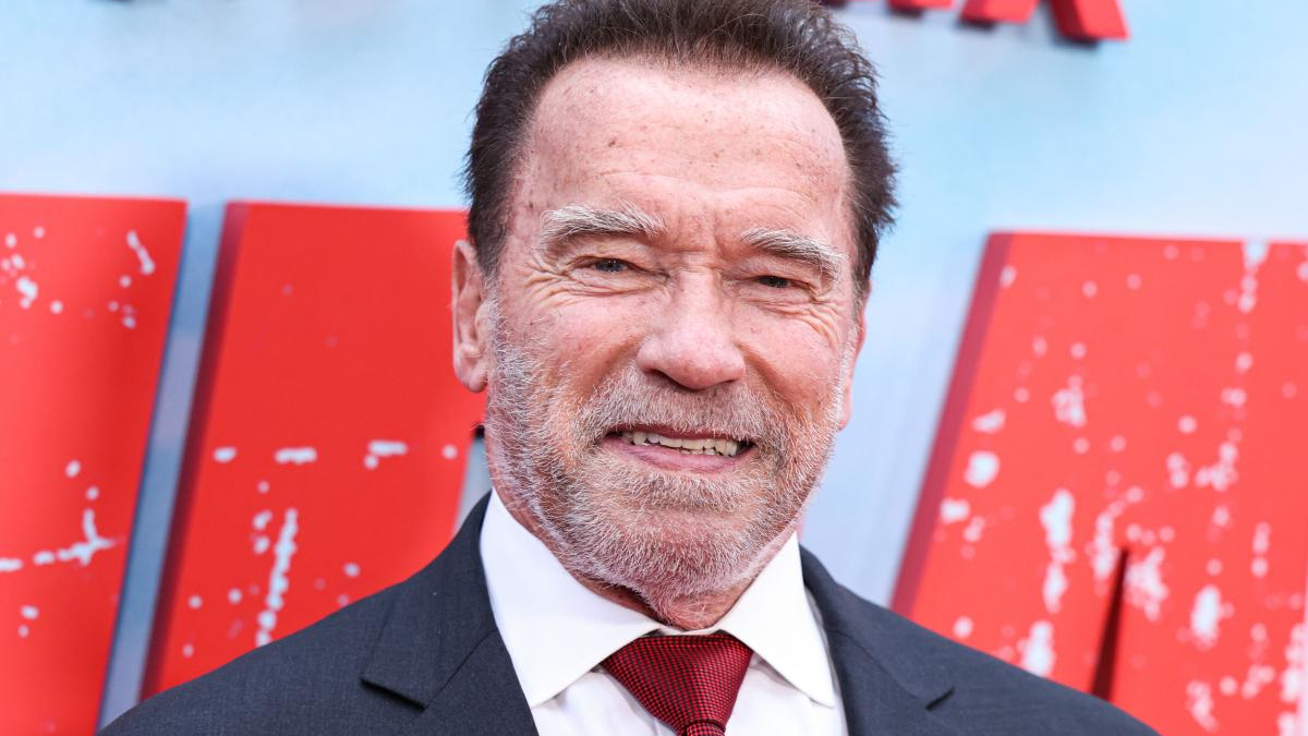 Smiling man in a dark suit and white shirt with a red tie, standing in front of a red backdrop.