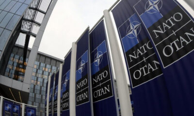 Row of dark blue NATO banners with the emblem and 'NATO' along a modern glass building exterior.