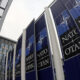 Row of dark blue NATO banners with the emblem and 'NATO' along a modern glass building exterior.