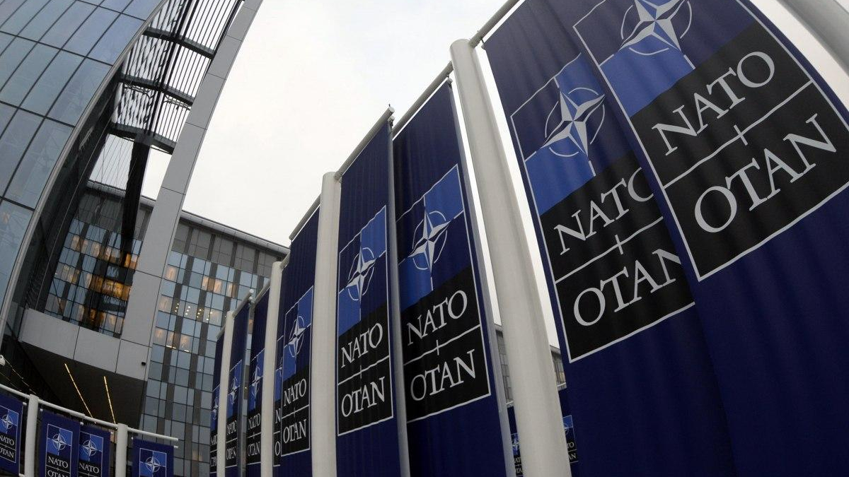 Row of dark blue NATO banners with the emblem and 'NATO' along a modern glass building exterior.
