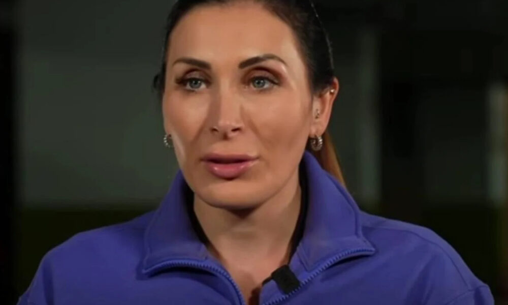 Woman in a blue jacket wearing a small microphone speaks directly to the camera in a dim setting.