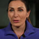 Woman in a blue jacket wearing a small microphone speaks directly to the camera in a dim setting.