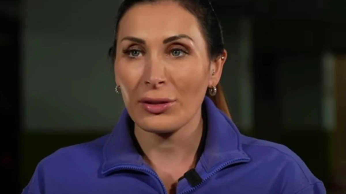 Woman in a blue jacket wearing a small microphone speaks directly to the camera in a dim setting.