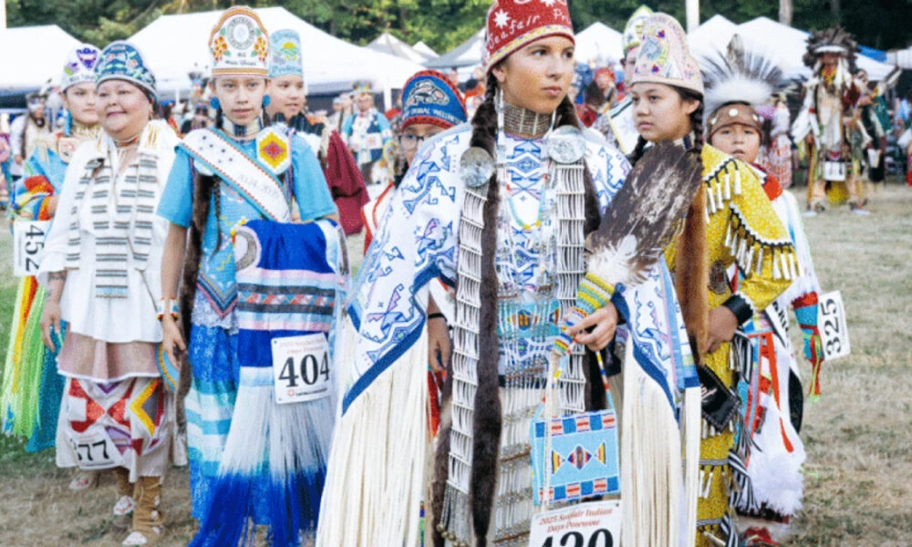 Group of Indigenous dancers in colorful ceremonial regalia at an outdoor festival, wearing numbered bibs.