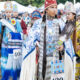 Group of Indigenous dancers in colorful ceremonial regalia at an outdoor festival, wearing numbered bibs.