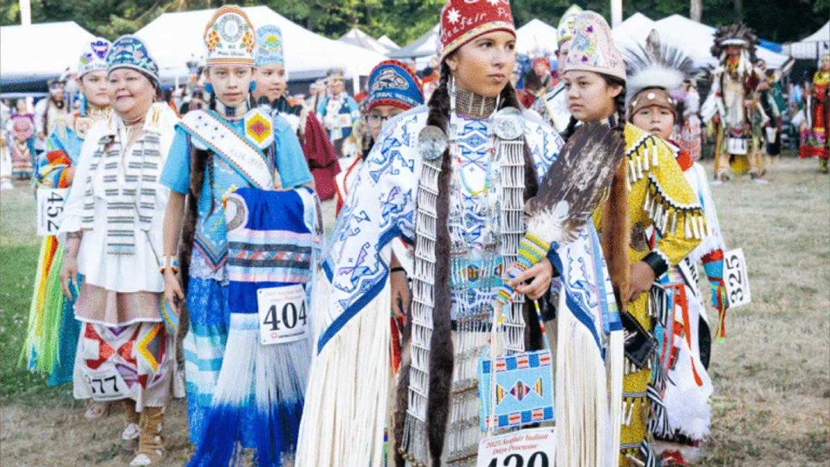 Group of Indigenous dancers in colorful ceremonial regalia at an outdoor festival, wearing numbered bibs.