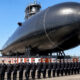 A row of sailors in dress uniforms standing at attention in front of a huge black submarine on a sunny tarmac.
