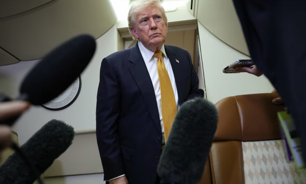 Man in a dark suit with a yellow tie stands in an airplane cabin as reporters with large microphones surround him.