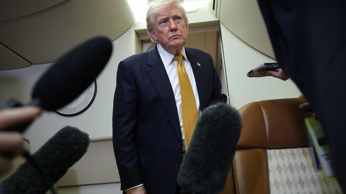 Man in a dark suit with a yellow tie stands in an airplane cabin as reporters with large microphones surround him.