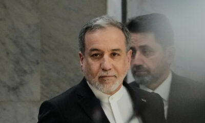 Middle-aged man with gray beard in a dark suit and white shirt looking at the camera, with a soft reflection in glass nearby.