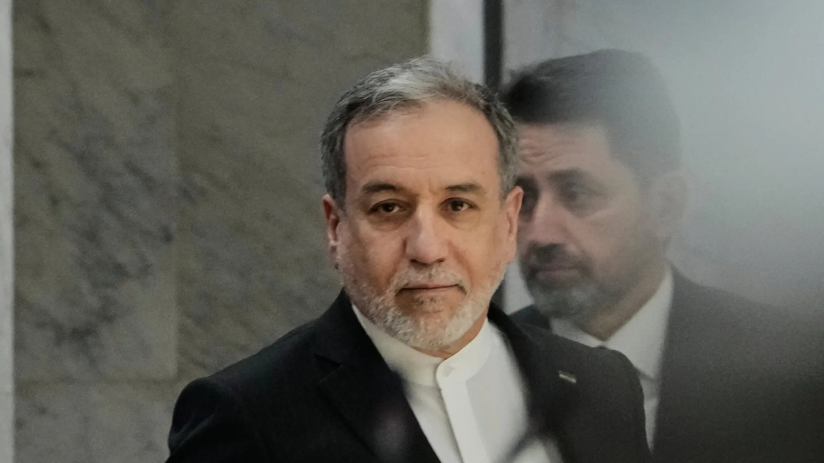 Middle-aged man with gray beard in a dark suit and white shirt looking at the camera, with a soft reflection in glass nearby.