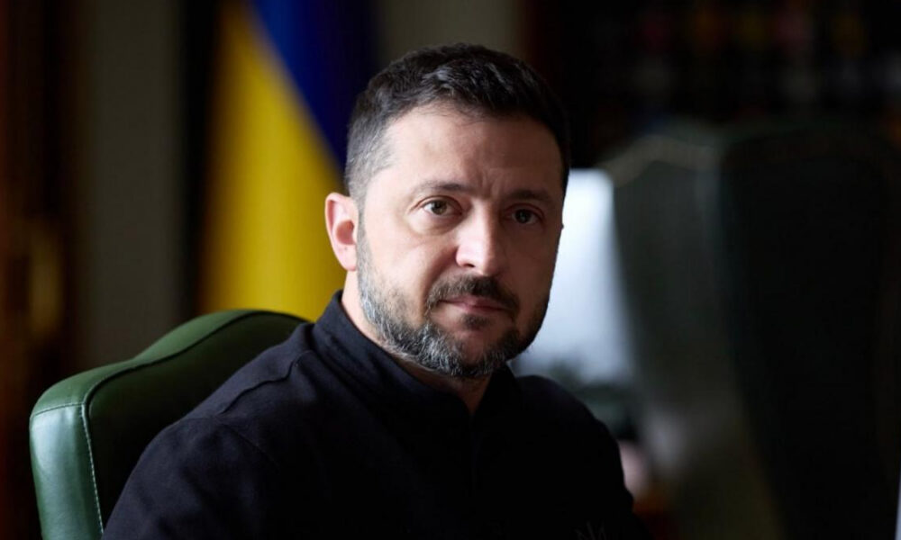 Man with short dark hair and beard sits in a green chair, wearing a black turtleneck, with a blue-yellow flag in the background.