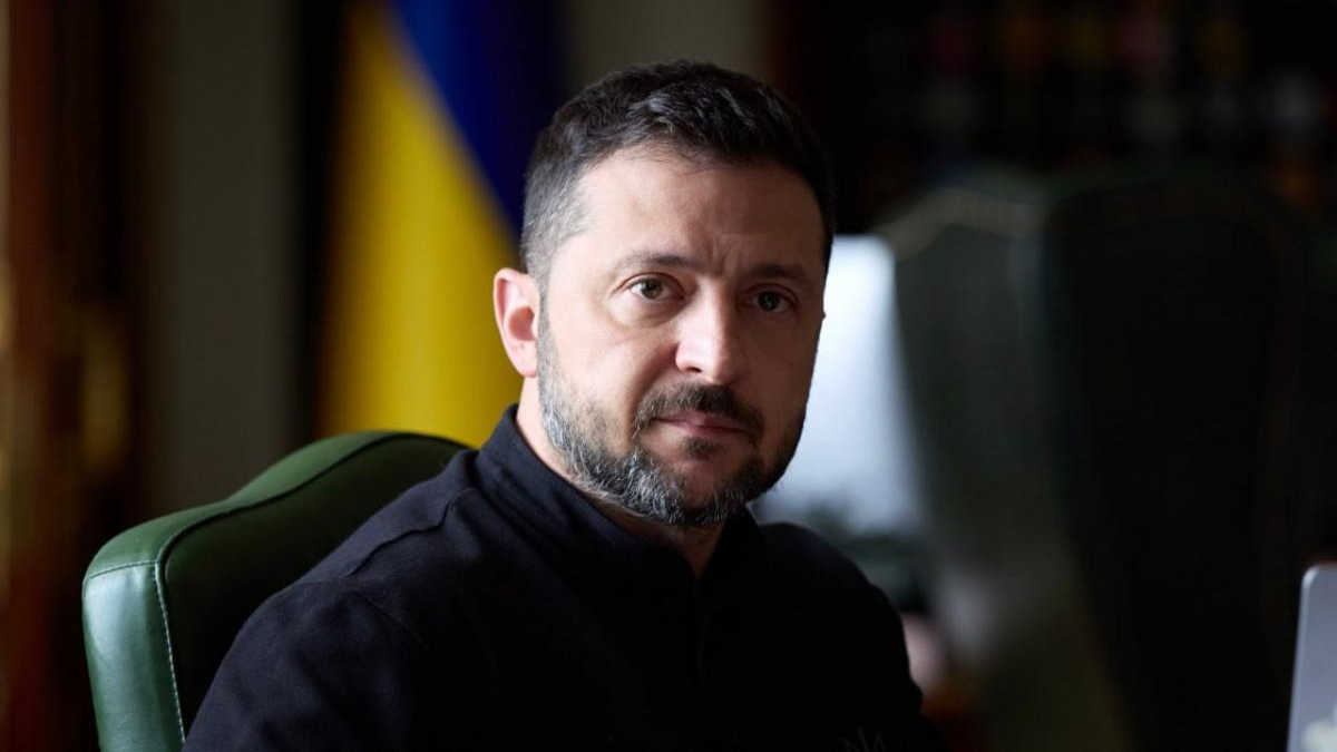 Man with short dark hair and beard sits in a green chair, wearing a black turtleneck, with a blue-yellow flag in the background.