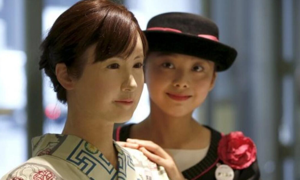 Two women posing indoors: a woman in a patterned kimono stands at the front while a friend in a black hat with a pink ribbon and flower smiles behind her and rests a hand on her shoulder.