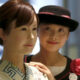Two women posing indoors: a woman in a patterned kimono stands at the front while a friend in a black hat with a pink ribbon and flower smiles behind her and rests a hand on her shoulder.