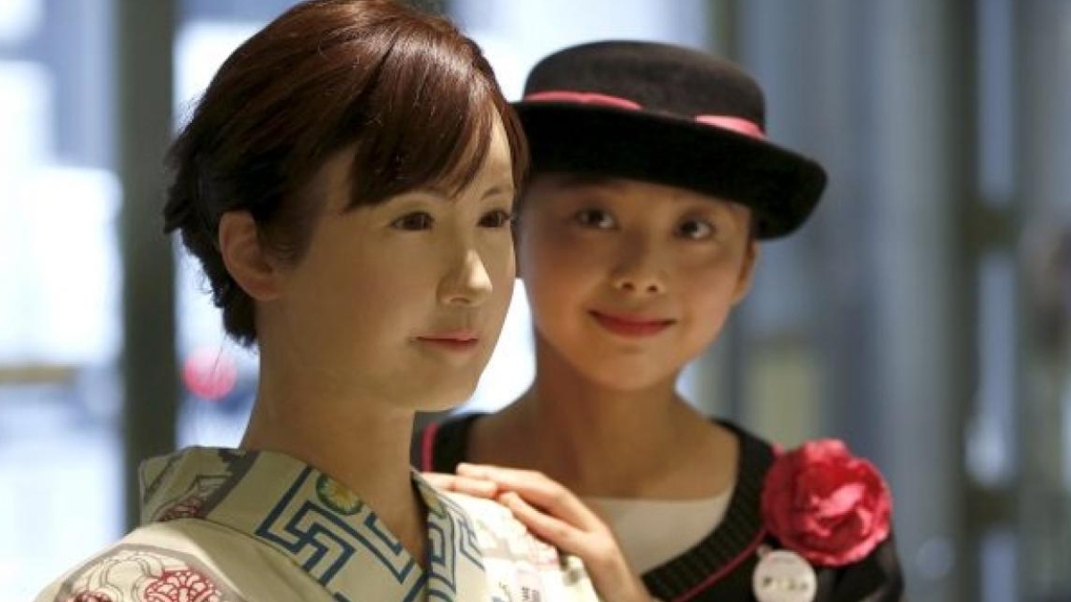 Two women posing indoors: a woman in a patterned kimono stands at the front while a friend in a black hat with a pink ribbon and flower smiles behind her and rests a hand on her shoulder.