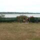 People walk along a grassy area beside a lake, with a green fence and shrubbery in the background and a distant city across the water.