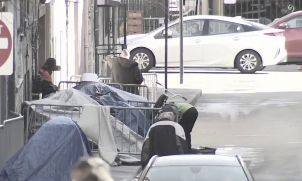 Urban sidewalk with makeshift tents and people sitting among barricades beside parked cars.