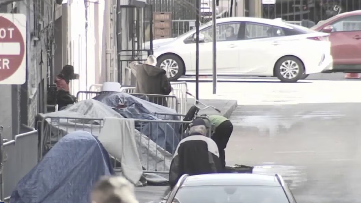 Urban sidewalk with makeshift tents and people sitting among barricades beside parked cars.