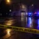 Night street at night with police cars flashing blue and red lights; yellow 'CRIME SCENE DO NOT CROSS' tape across the wet road and cordoned area in the foreground.
