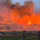 Wildfire burning across a grassy hillside at dusk, with flames and smoke rising behind a wooden fence in a rural field.