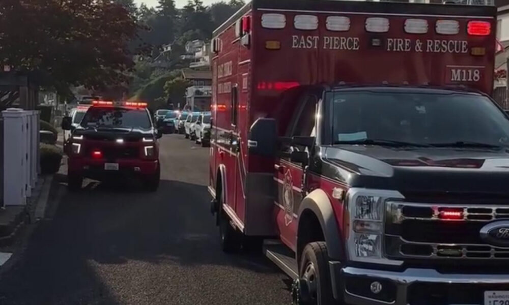 Front-right view of a red East Pierce Fire & Rescue ambulance with flashing lights, parked on a suburban street with other emergency vehicles in the background.