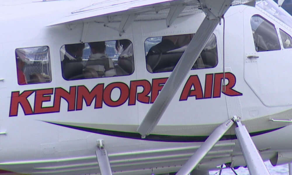 Side view of a white helicopter with bold red 'KENMORE AIR' branding along the fuselage, cockpit windows visible and landing gear struts in the foreground.