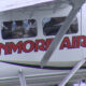 Side view of a white helicopter with bold red 'KENMORE AIR' branding along the fuselage, cockpit windows visible and landing gear struts in the foreground.