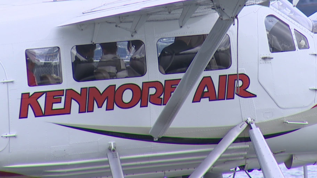 Side view of a white helicopter with bold red 'KENMORE AIR' branding along the fuselage, cockpit windows visible and landing gear struts in the foreground.