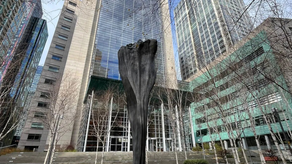 Abstract black sculpture rising from the plaza in front of a glass skyscraper, with leafless trees and steps leading to the entrance.