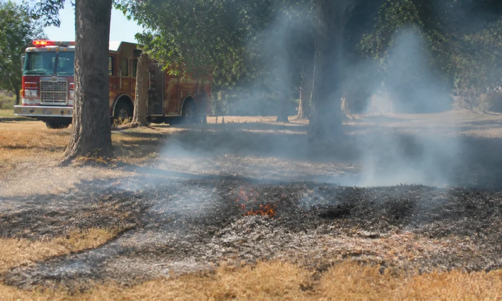 Fire truck parked on a grassy field while smoke rises from a small brush fire in the foreground.