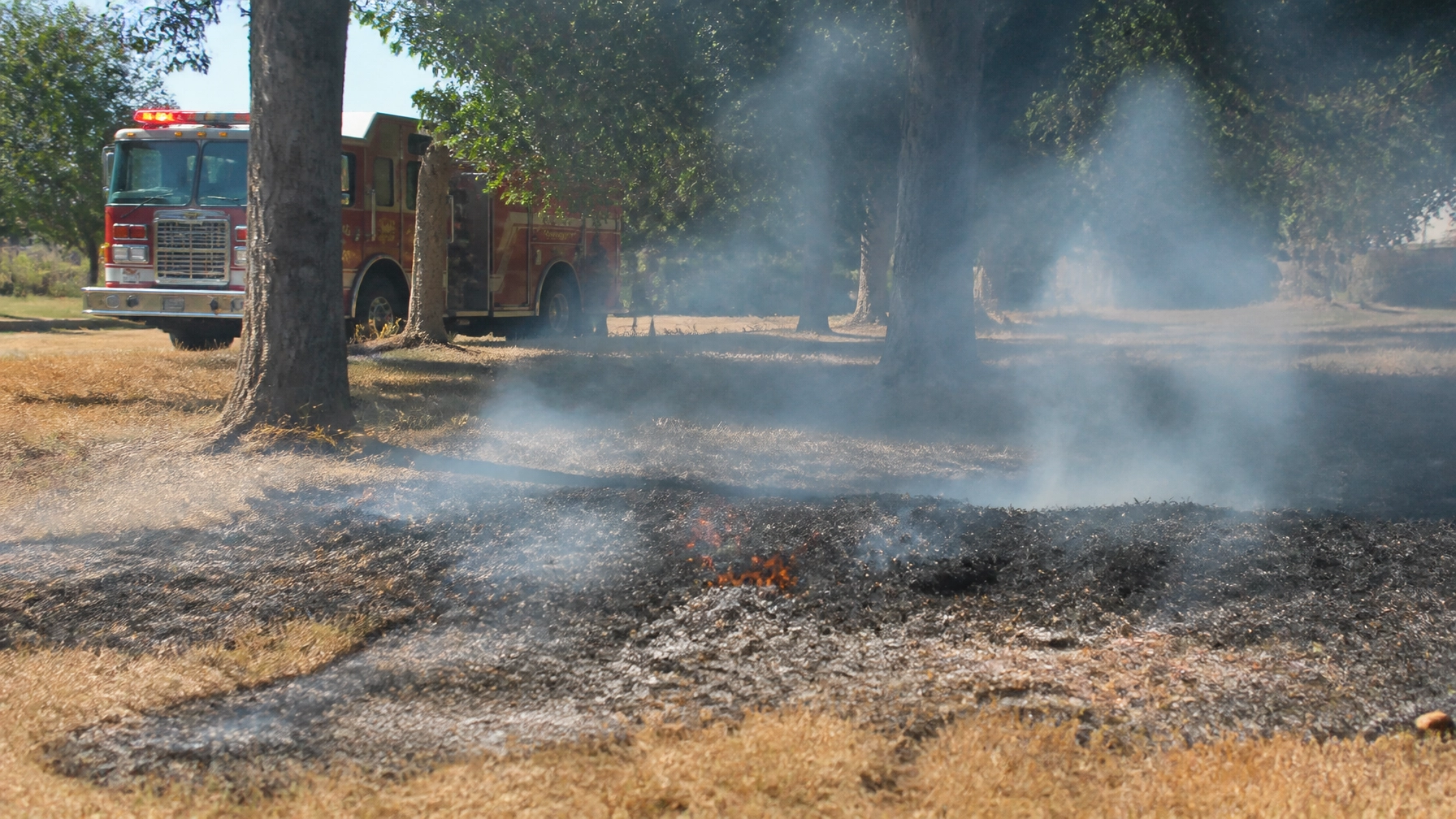 Fire truck parked on a grassy field while smoke rises from a small brush fire in the foreground.