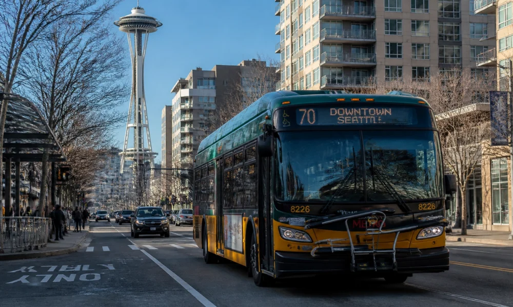 Green and yellow Seattle city bus on a street with the Space Needle and modern apartment buildings in the background, route 70 to Downtown Seattle visible on the bus front.