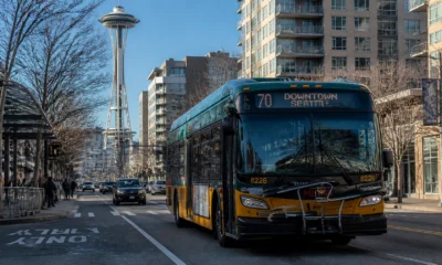 Green and yellow Seattle city bus on a street with the Space Needle and modern apartment buildings in the background, route 70 to Downtown Seattle visible on the bus front.