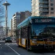 Green and yellow Seattle city bus on a street with the Space Needle and modern apartment buildings in the background, route 70 to Downtown Seattle visible on the bus front.