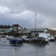 Shopping plaza with a row of stores, a wet parking lot, and several cars; an American flag on a tall pole stands near the center under gray clouds.