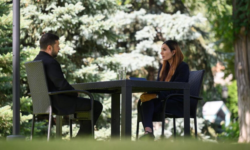 Two professionals seated at a wicker table outdoors, facing each other in a garden setting with glasses on the table, mid-conversation in a sunny yard.