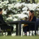 Two professionals seated at a wicker table outdoors, facing each other in a garden setting with glasses on the table, mid-conversation in a sunny yard.