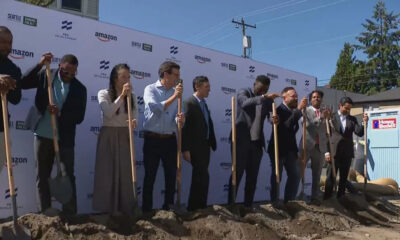 Group of diverse professionals at a groundbreaking ceremony, each holding a shovel and standing in dirt in front of a branded backdrop with Amazon logos.