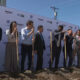 Group of diverse professionals at a groundbreaking ceremony, each holding a shovel and standing in dirt in front of a branded backdrop with Amazon logos.