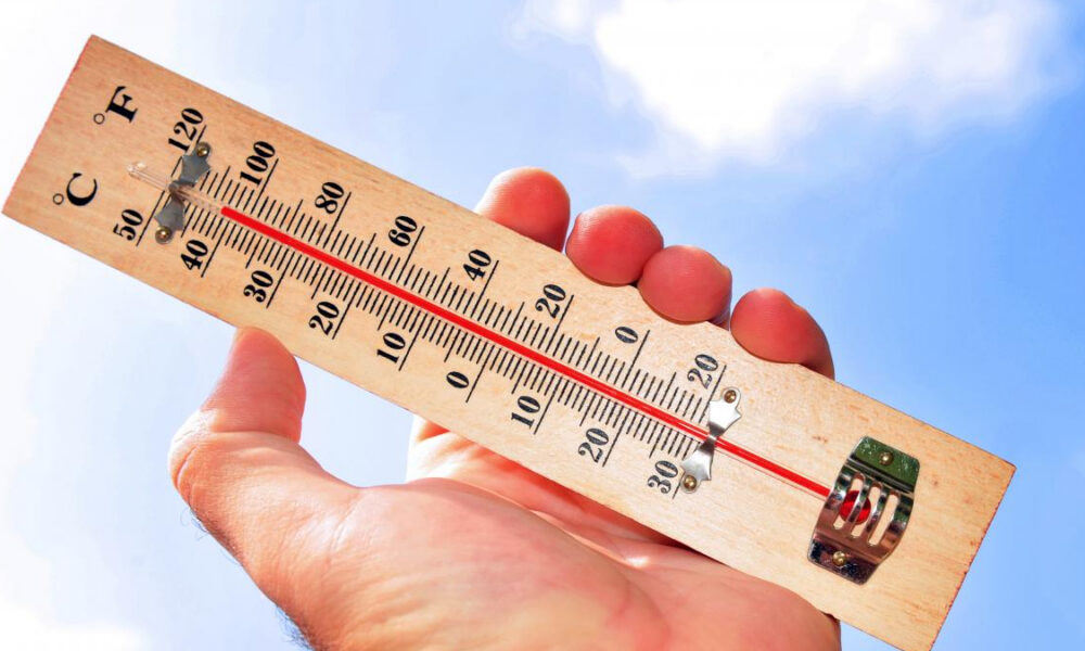 Hand holding a wooden thermometer up to a clear blue sky, showing the temperature with a red liquid line.